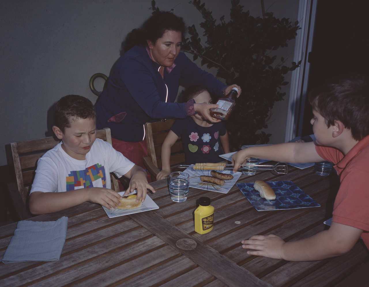 John about to eat his hot dog, Jackie opening hte ketchup for Sophia, and James grabbing a wiener with the tongs at Jules and Elly's house