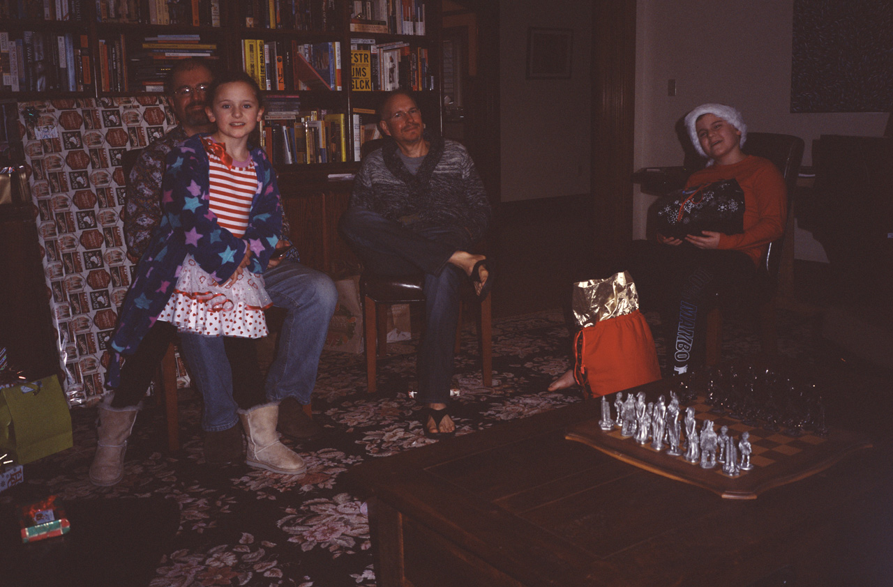 Sophia sitting on Steve's lap, Marc looking amused, and James waiting impatiently to open his next gift, in the Piano Room on Christmas morning