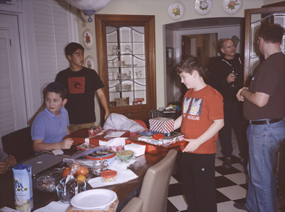 John looking to his right, Mikhil standing behind John, James holding his Birthday gifts, Marc reaching in the glass cabinet for a wine glass, and Steve looking at Marc, in the kitchen