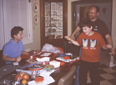 John sitting at the kitchen dining table ready to open his Birthday gifts, as Steve helps James sit down at hte table with his gifts