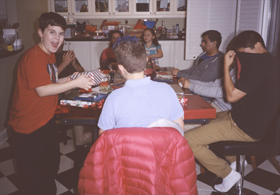 James standing and looking surprised, Steve clapping behind James, John, sitting at the kitchen table with his back to the camera, Jackie and Sophia across the table from John, Naresh looking at his laptop, and Mikhil covering his eyes with his shirt, in the kitchen