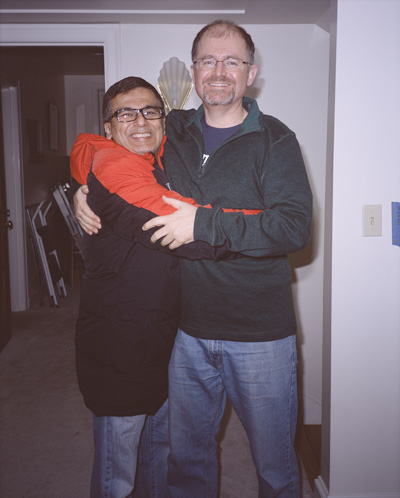 Naresh and Steve hugging and standing in the kitchen, with the hall and Jackie's office behind them